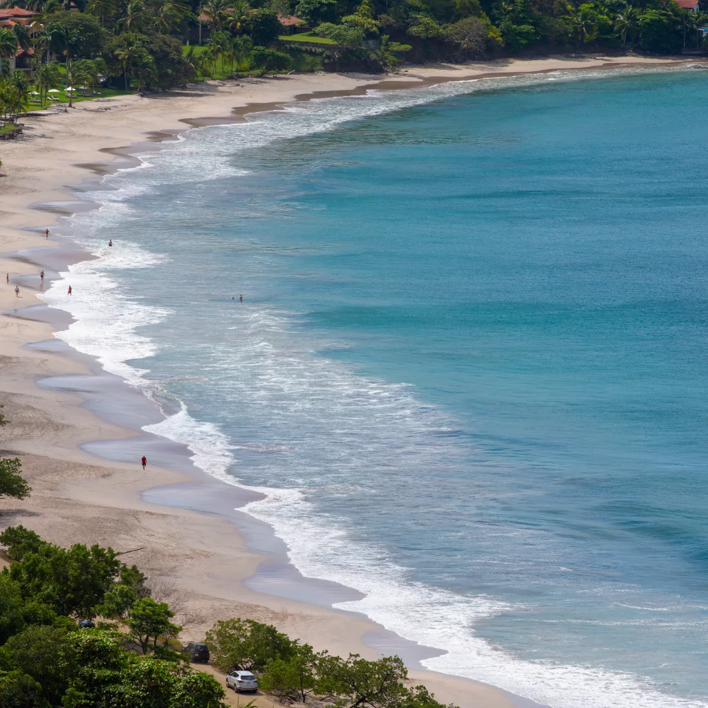 Aerial view of Playa Flamingo's white sand beach and turquoise waters, with lush tropical vegetation and a handful of visitors along the shoreline, Guanacaste, Costa Rica