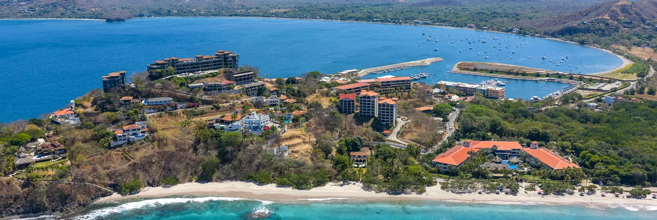 Aerial view of Playa Flamingo showing the white sand beach, turquoise waters, residential and resort buildings on the hillside, and Marina Flamingo in the background, Guanacaste, Costa Rica