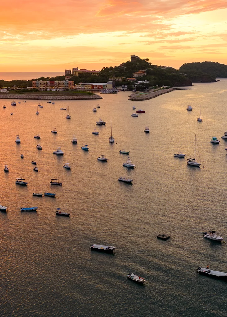 Aerial view of Marina Flamingo at sunset with dozens of boats anchored in the golden bay, the colorful marina buildings and hillside community visible in the background, Guanacaste, Costa Rica