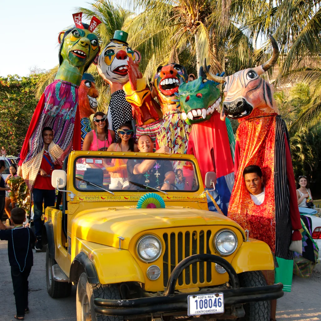 Community members celebrating a Costa Rican cultural festival in Playa Flamingo, riding in a decorated yellow jeep surrounded by colorful giant puppet figures and masks, Guanacaste, Costa Rica