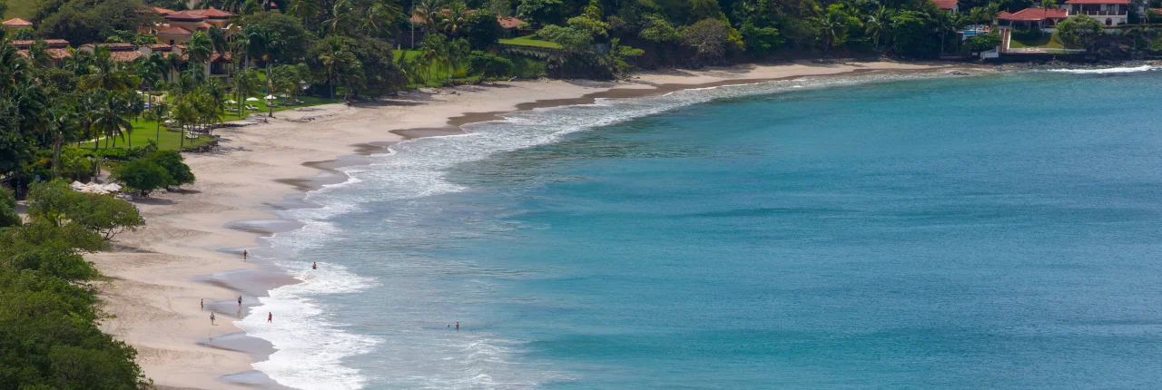 Aerial view of Playa Flamingo's curved white sand beach with turquoise waters, lush tropical vegetation, and luxury residences along the shoreline, Guanacaste, Costa Rica