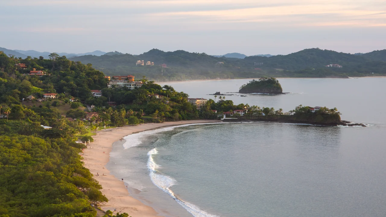Aerial drone view of Playa Flamingo at dusk, showing the curved white sand beach, calm bay waters, lush tropical hillside, and coastal homes, Guanacaste, Costa Rica