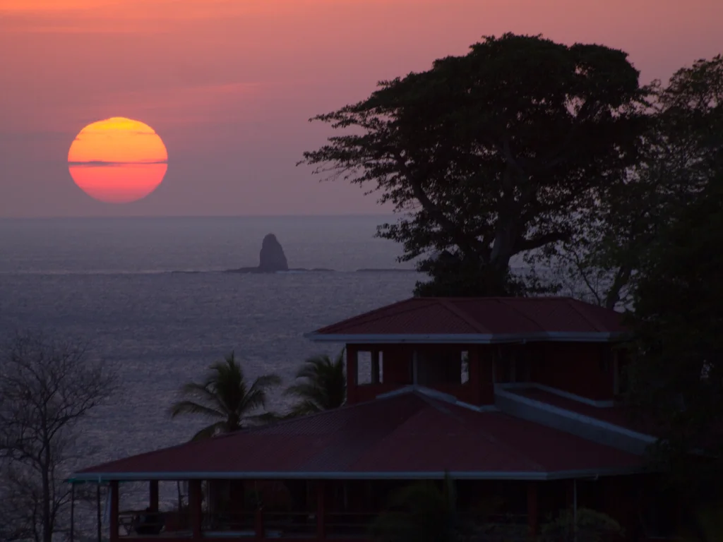 A vivid red and orange sunset over the Pacific Ocean viewed from Playa Flamingo, with a coastal home, tropical trees, and a rocky island silhouetted against the glowing sky, Guanacaste, Costa Rica
