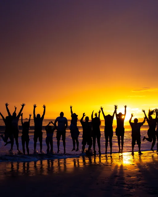 Silhouettes of a large group of people celebrating on the beach at sunset, arms raised, with golden orange sky reflecting on the wet sand, Playa Flamingo, Guanacaste, Costa Rica
