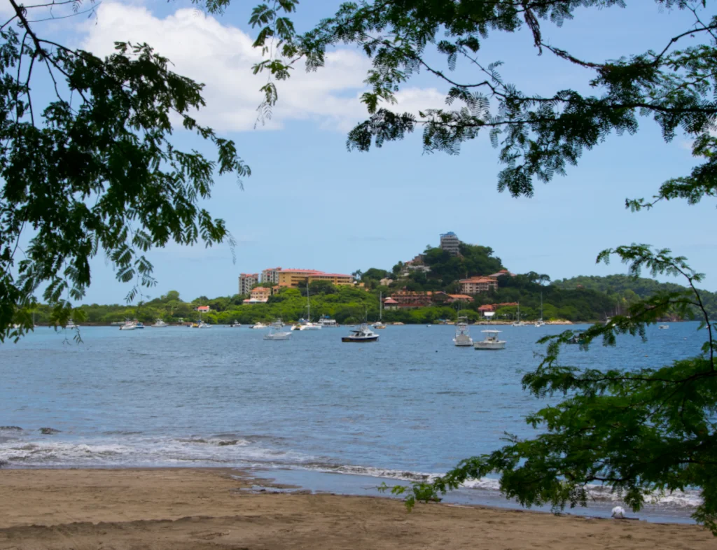 View of Playa Flamingo bay framed by tropical tree branches, with boats anchored in calm water and the hillside community and resort buildings visible in the background, Guanacaste, Costa Rica
